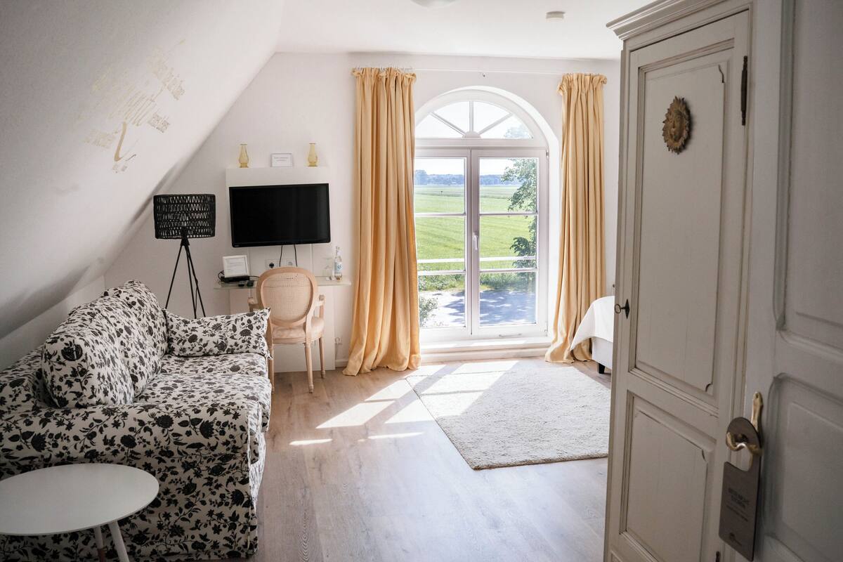 Ein Zimmer mit einer Couch, einem kleinen Tisch, einem Fernseher, einem Sessel und einer Tür, die auf einen Balkon mit Vorhängen führt.