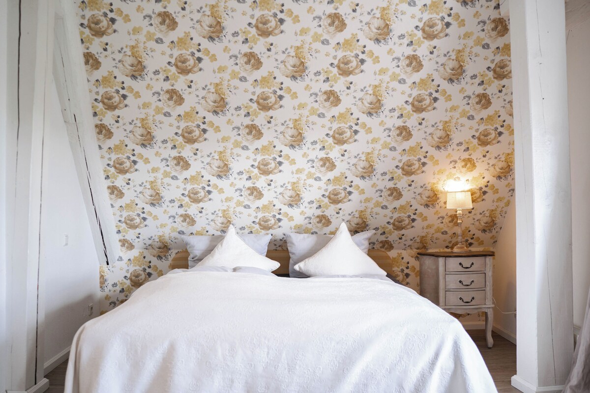 Un dormitorio con papel tapiz floral, una cama con ropa de cama blanca y una mesita de noche con una lámpara.