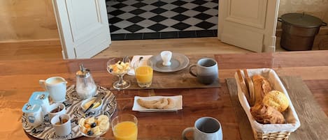 Ein Frühstückstisch mit verschiedenen Speisen und Getränken, ein karierter Boden und Blick in einen Raum mit einem Bett und einer Pflanze.