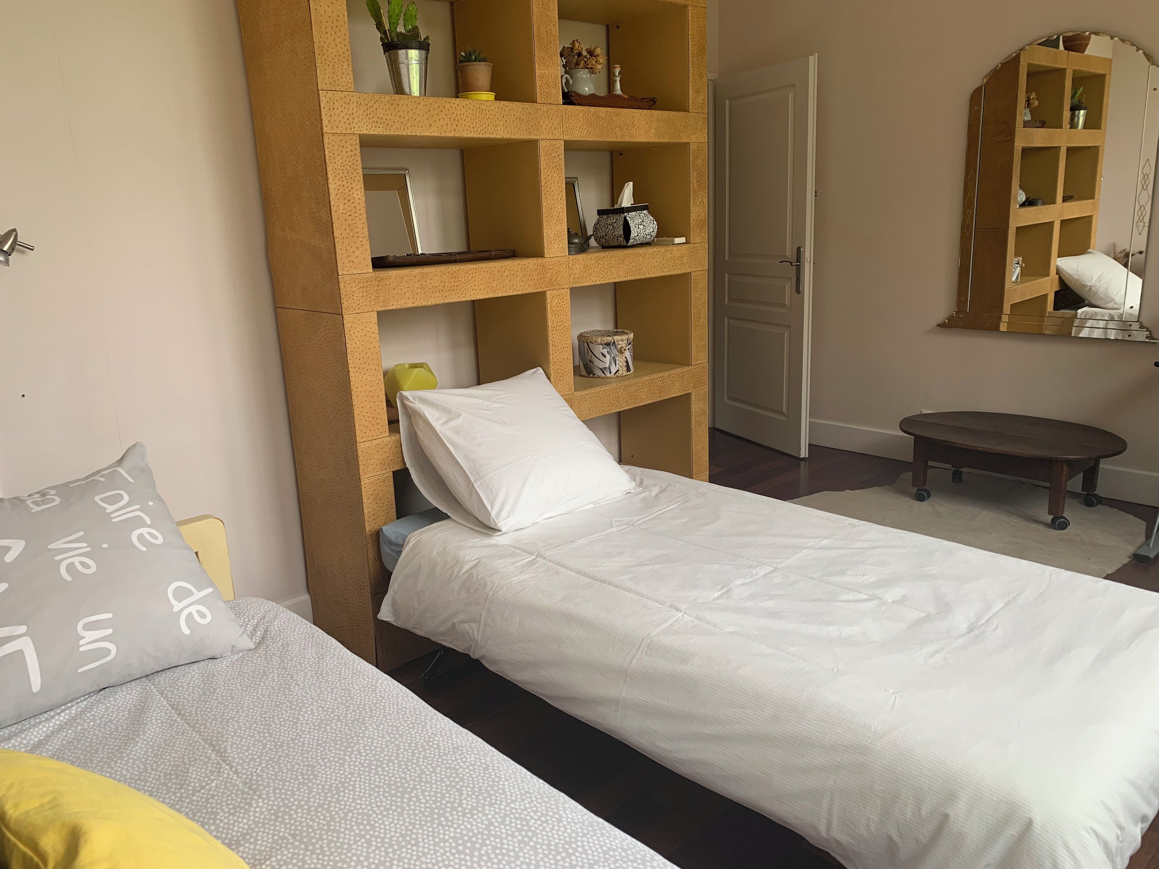 A bedroom with two beds, a wooden shelf unit with various items, a small round table, and a mirror on the wall.