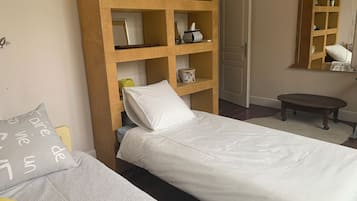 A bedroom with two beds, a wooden shelf unit with various items, a small round table, and a mirror on the wall.