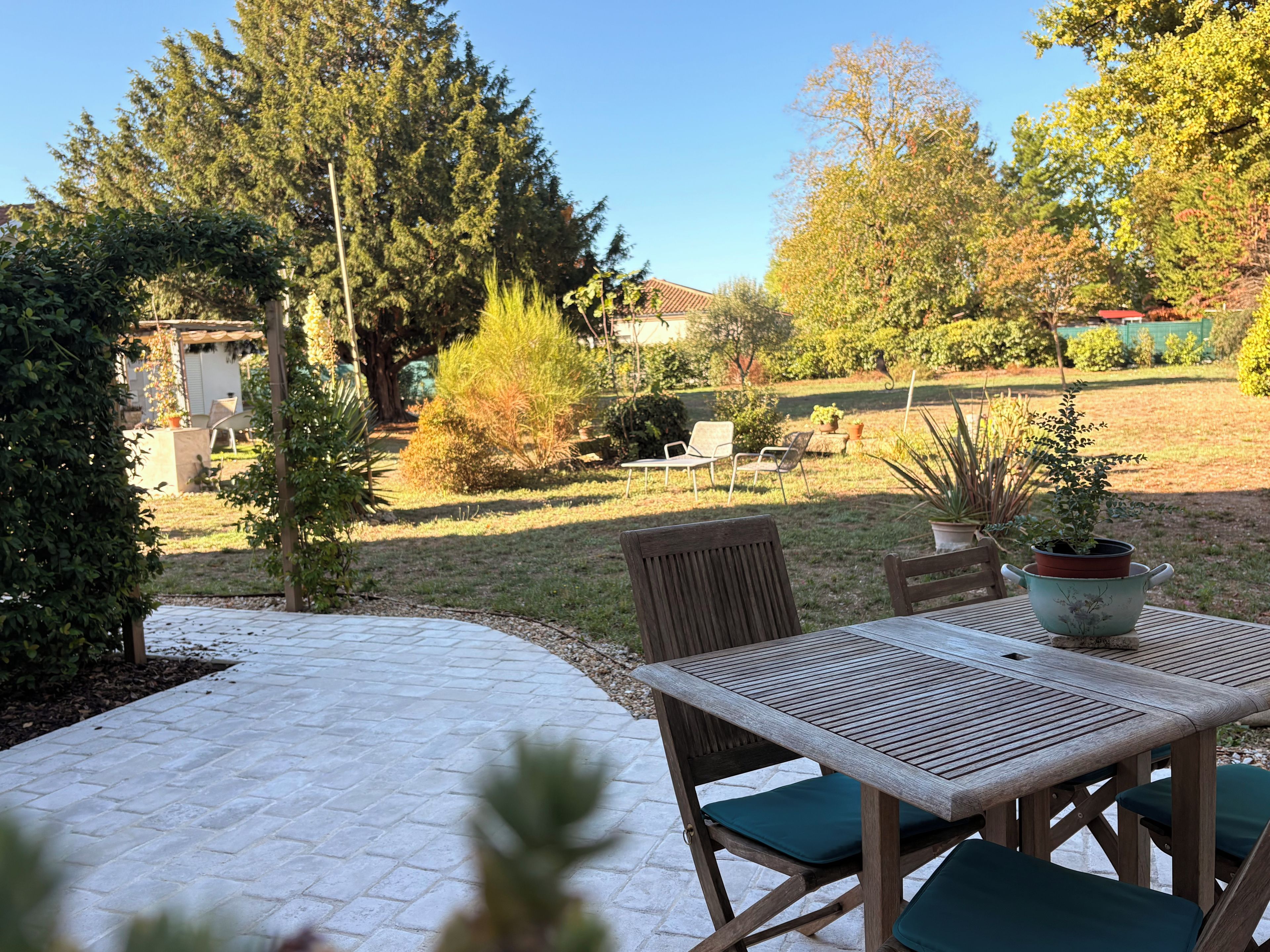 An outdoor patio with a table and chairs, surrounded by a garden with various plants and trees.