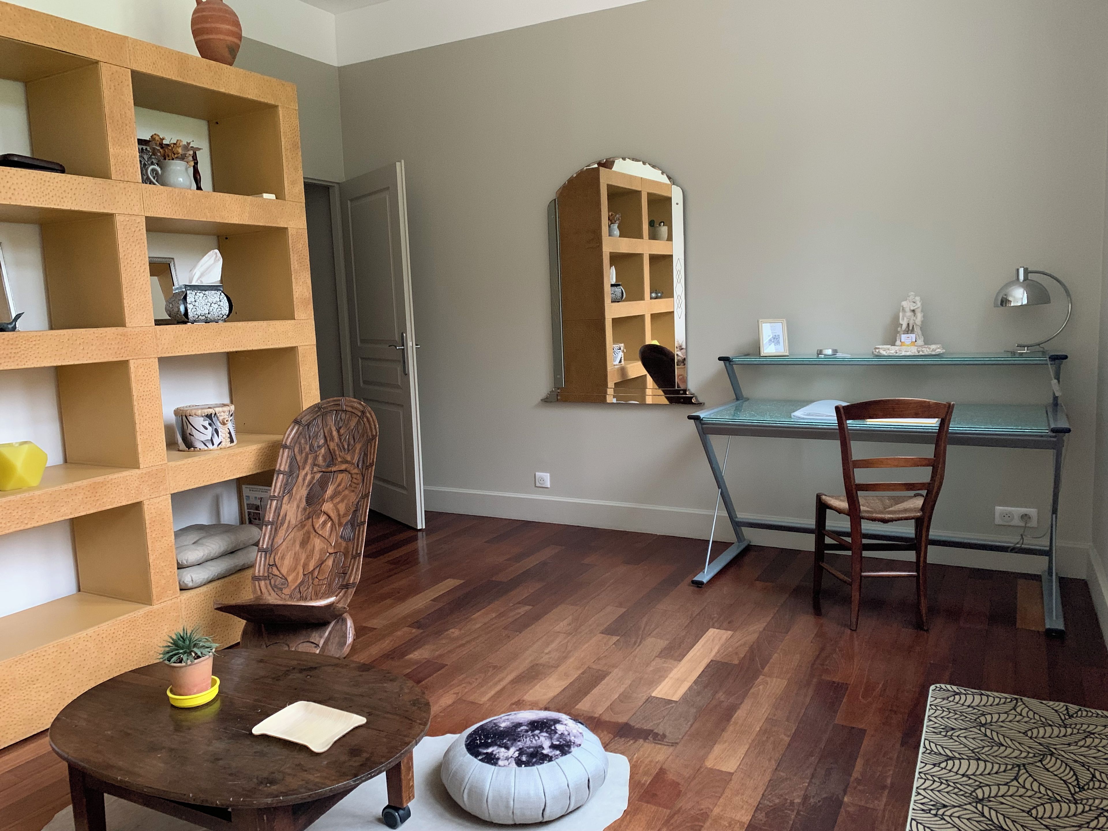 Une pièce avec une bibliothèque en bois, un bureau avec une chaise, une table ronde avec un coussin et un miroir accroché au mur.