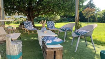 An outdoor seating area with wooden tables and chairs, a large tree, and a grassy lawn.