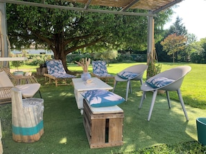 An outdoor seating area with wooden tables and chairs, a large tree, and a grassy lawn.