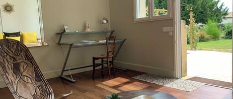 A room with a wooden chair, a desk with a computer, a small table with a plant, and a view of the outdoors through a window.