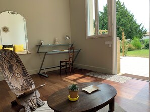 Una habitación con una silla de madera, un escritorio con una computadora, una mesita con una planta y vistas al exterior a través de una ventana.
