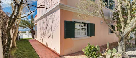 A two-story building with a pink facade, green shutters, and a paved pathway leading to the entrance.