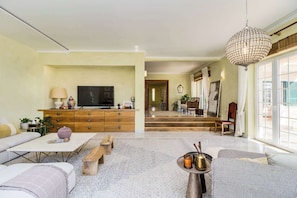 A living room with a TV, wooden furniture, a chandelier, and a view of the dining area. - Luxury Coastal Villa with Heated Pool,  Basketball Court & Ocean Views near Lisbon (Atalaia de Cima)