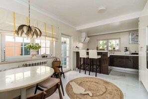 A modern kitchen with a dining area, featuring a round table, bar stools, and a large window. - Luxury Coastal Villa with Heated Pool,  Basketball Court & Ocean Views near Lisbon (Atalaia de Cima)