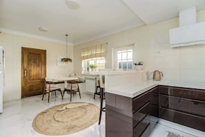 A modern kitchen with a dining area, white tiles, and a wooden door. - Luxury Coastal Villa with Heated Pool,  Basketball Court & Ocean Views near Lisbon (Atalaia de Cima)