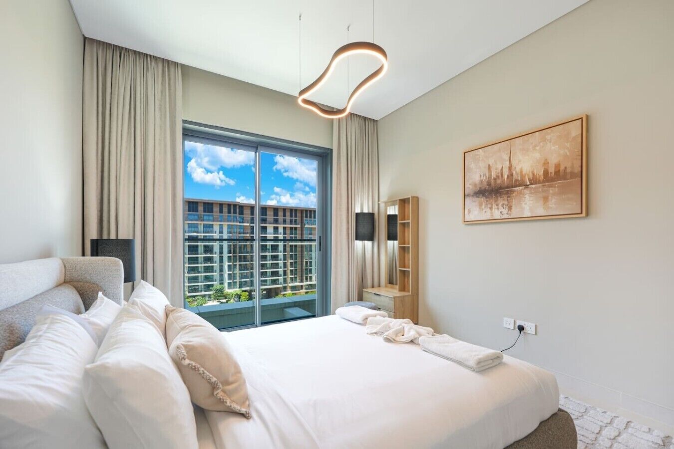 Quarto de hotel com uma cama grande, vista de edifícios pela janela, um quadro na parede e uma luminária moderna.