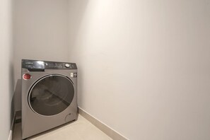 Eine kompakte, moderne Waschmaschine steht in einer Ecke des Raumes, dessen Wand schlicht gehalten und der Boden hell ist.