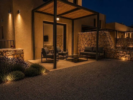 A modern outdoor seating area with stone walls and a pergola, surrounded by a gravel driveway.. Elafonisos Village Boutique Residences