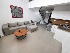 Un espacio de estar moderno con un sofá seccional gris, una mesa de centro de madera, una pequeña mesa redonda blanca y una cocina con armarios blancos y encimera de madera.
