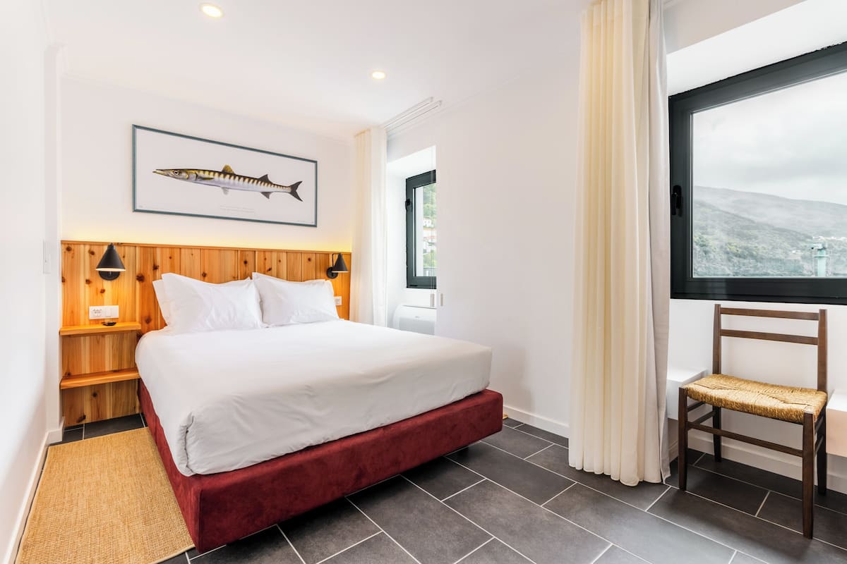 Habitación de hotel con cama, cabecera de madera, un cuadro con una imagen de peces, una silla y ventana con vistas.