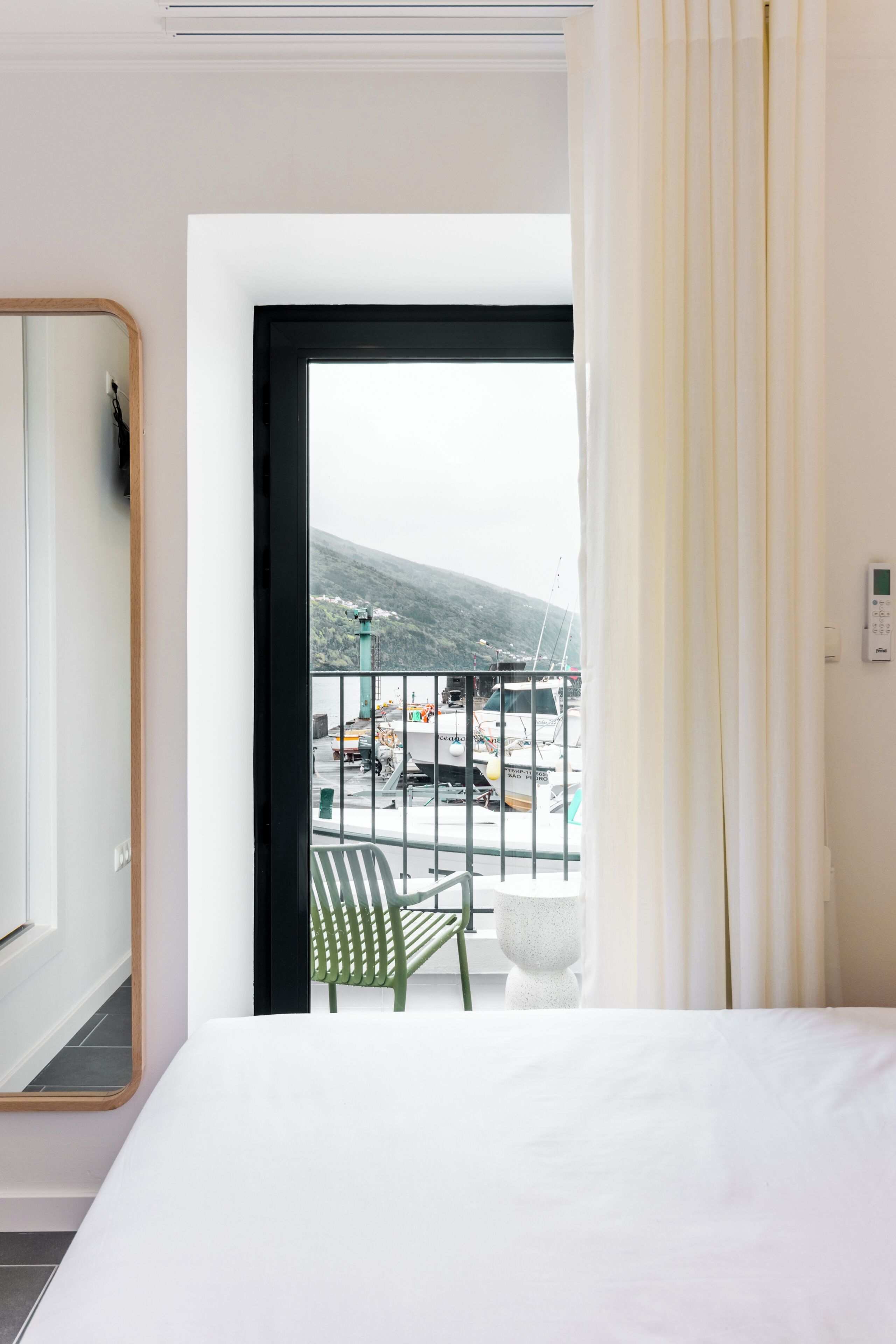 Habitación de hotel con cama, una silla verde, ventana con cortinas y vista a barcos y una montaña.