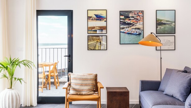 Een moderne woonkamer met een rieten plafondlamp, een bank, een stoel, een bijzettafeltje en uitzicht op de zee door een deur.