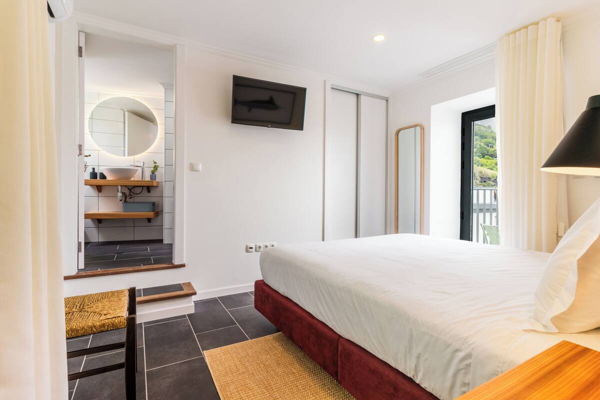 Un dormitorio con cama, televisor, espejo, ventana y baño con lavamanos y espejo.