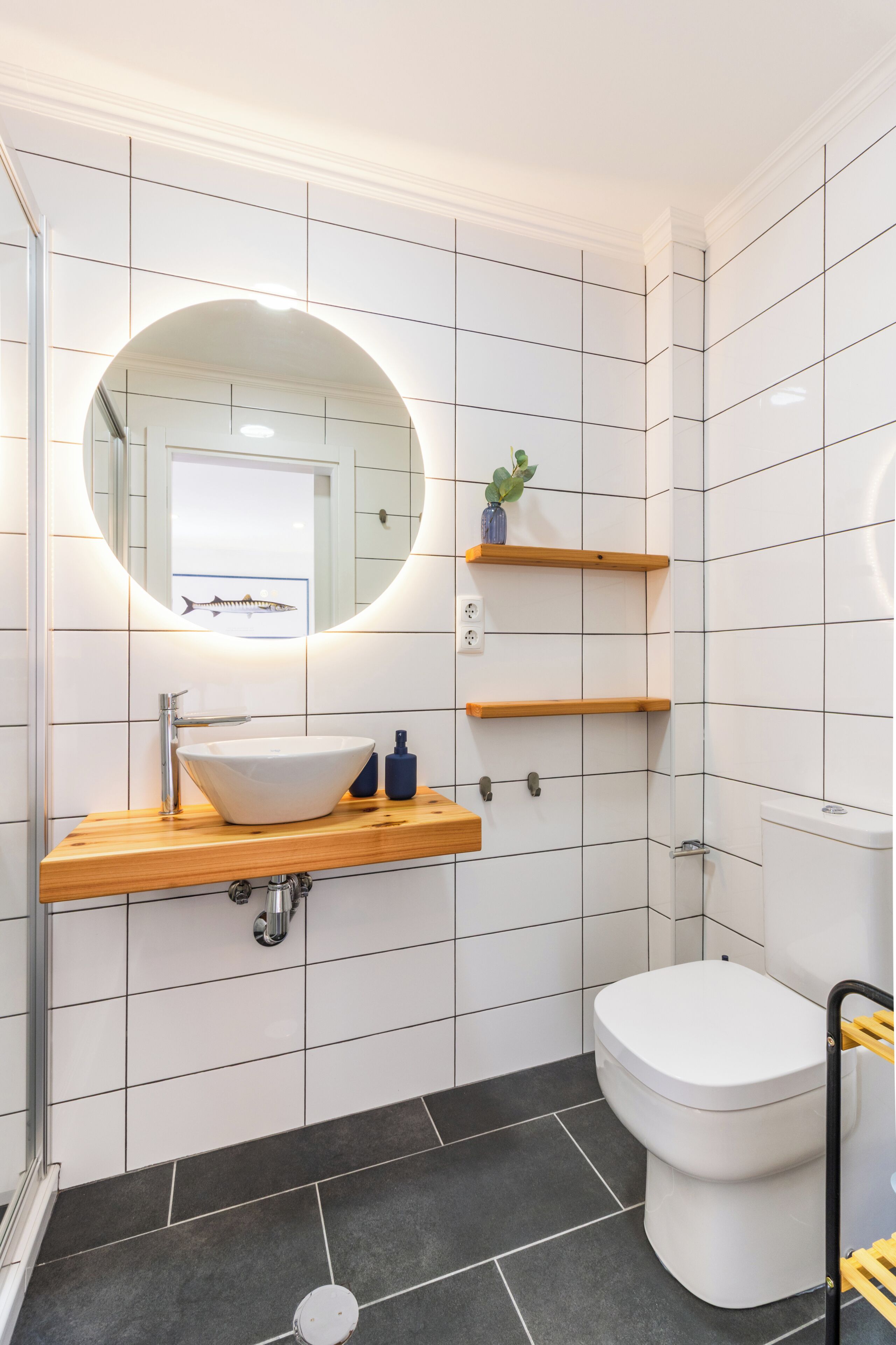 Un baño con un lavabo blanco, un espejo redondo y un inodoro. Hay estantes de madera sobre el lavabo y una planta pequeña en uno de ellos.