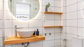 Un baño con un lavabo blanco, un espejo redondo y un inodoro. Hay estantes de madera sobre el lavabo y una planta pequeña en uno de ellos.