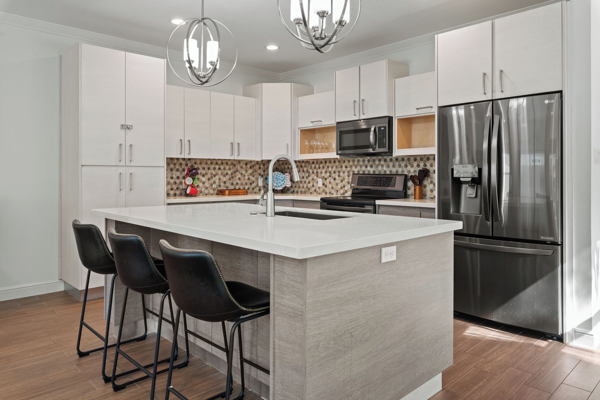 A modern kitchen with white cabinets, a stainless steel refrigerator, a microwave, and a large island with bar stools.