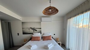 A modern hotel room with a large bed, two bedside tables, a wall-mounted air conditioner, and a large window with sheer curtains. - Studio Calypso 20 m from the sea (Mauguio)