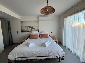 Ein modernes Hotelzimmer mit einem großen Bett, zwei Nachttischen, einer Wand-Klimaanlage und einem großen Fenster mit durchsichtigen Vorhängen.