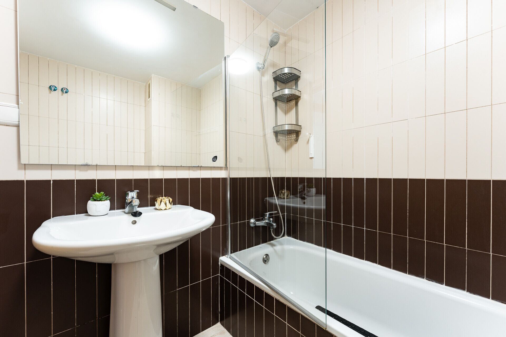 Ein Badezimmer mit einem weißen Waschbecken, einem Spiegel, einer Dusche mit Duschbrause und braunen Fliesenwänden.