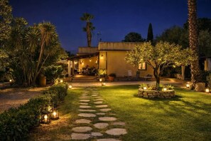 A well-lit outdoor area with a stone pathway leading to a building, surrounded by greenery and palm trees. - Donna Caterina House: Private Retreat in Central Sicily (Pietraperzia)