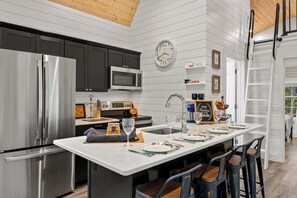 A modern kitchen with stainless steel appliances, a central island with bar stools, a clock, and a 'Welcome' sign. - Shenandoah, Firepit, peaceful Cabin w/salt water Hot tub! (Luray)