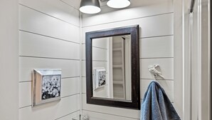 Un bagno con pareti bianche a pannelli, uno specchio con cornice scura, un appendi-asciugamani con un asciugamano e un quadro incorniciato appeso alla parete.