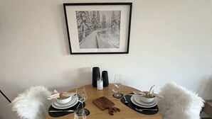 A round wooden dining table with a white fluffy chair, a framed picture on the wall, and a candle holder. - book2stay: Retro-Alpin Studio with balcony (Laax)