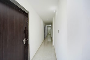 Un pasillo con una puerta de madera oscura a la izquierda, paredes blancas y suelo de baldosas.