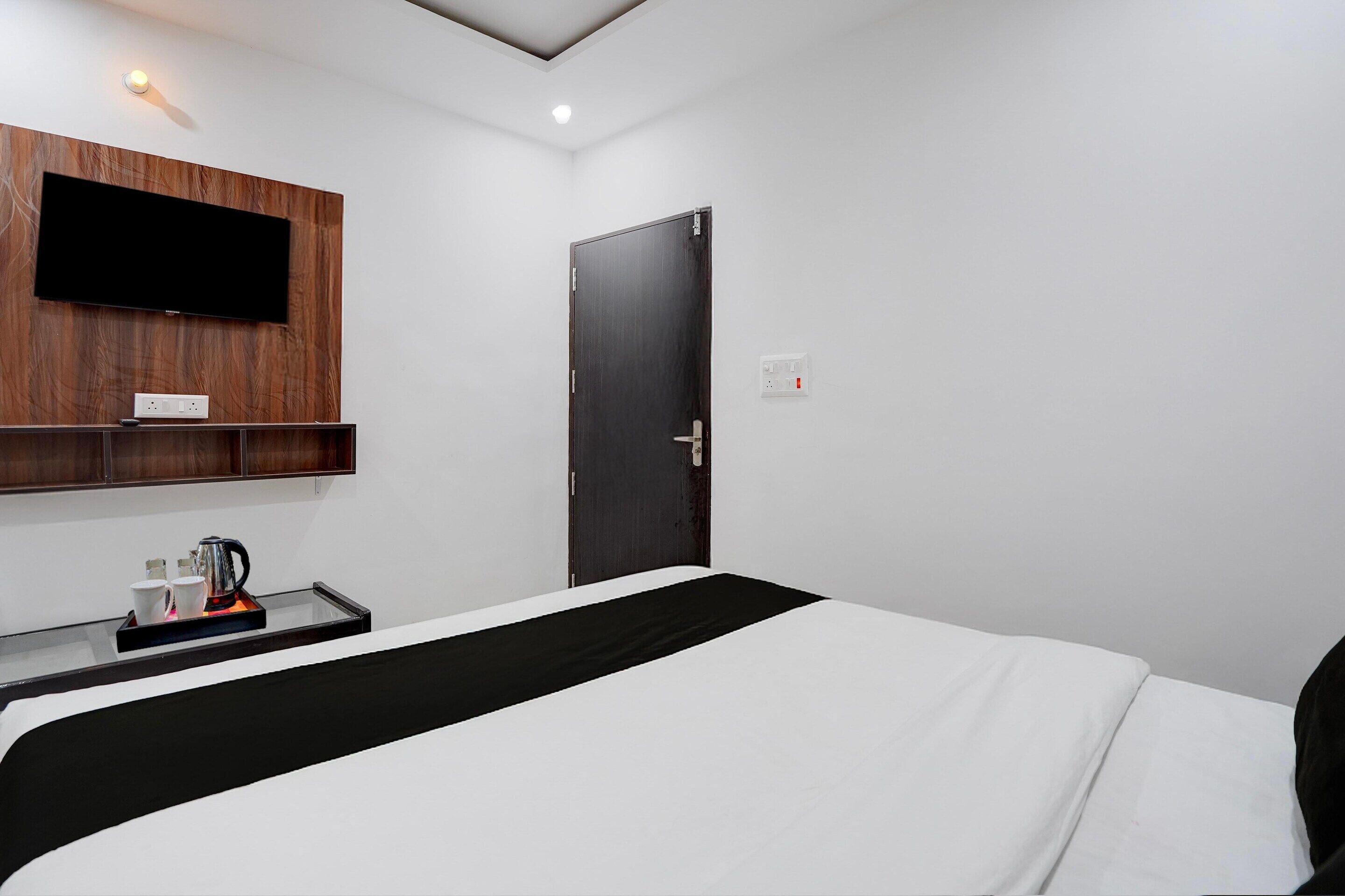 A hotel room with a bed, a TV mounted on a wooden panel, a bedside table with a kettle, and a door with a handle.