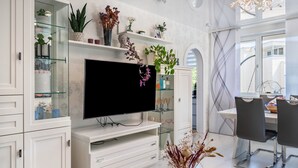 A modern living room with a flat-screen TV, white cabinetry, glass shelves, and a dining area with chairs.