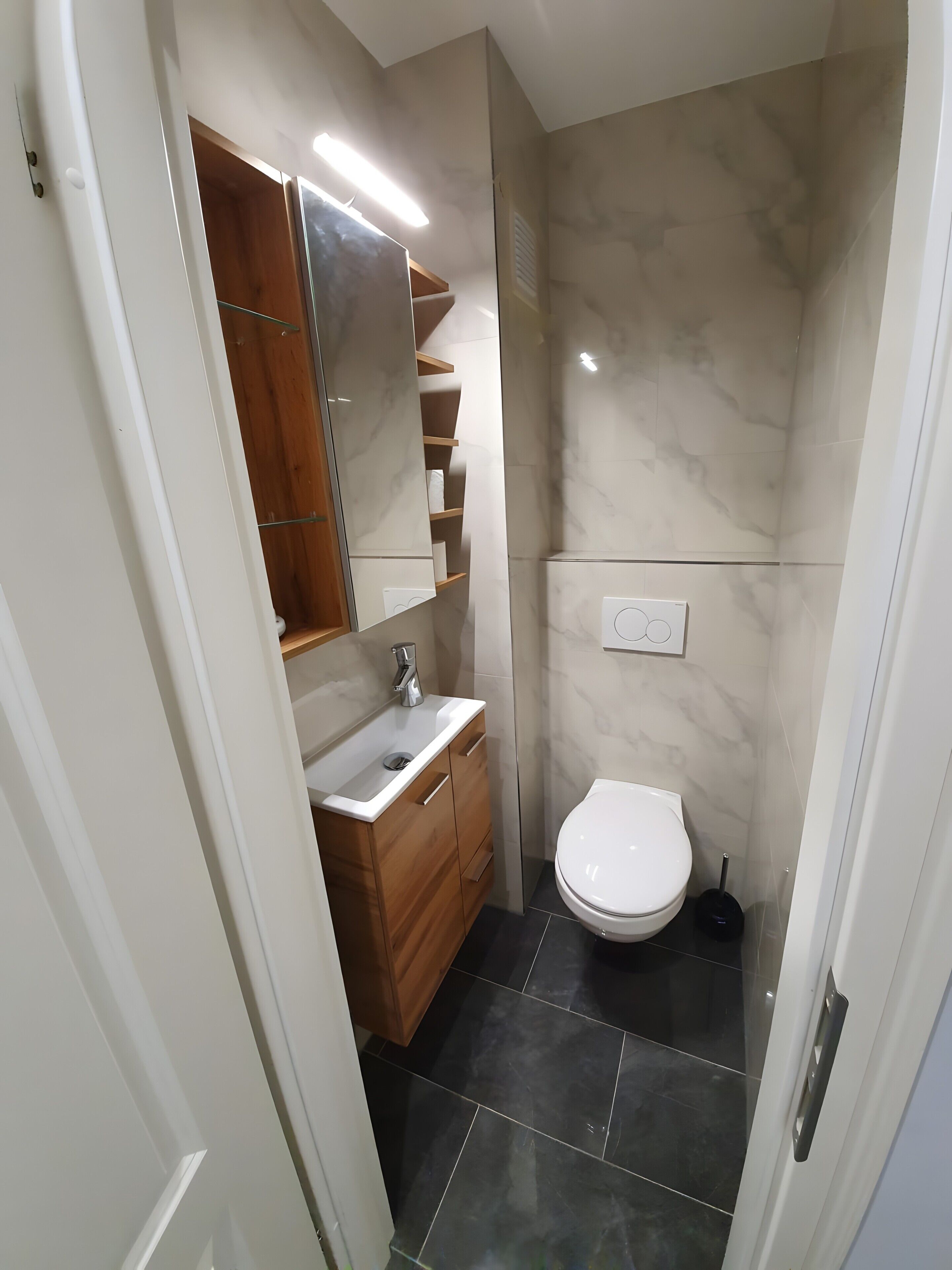 A compact bathroom with a white toilet, a wooden vanity with a mirror, and a marble wall.