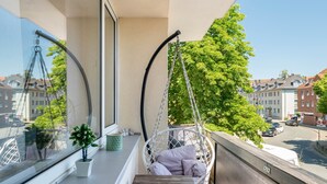 A balcony with a hanging chair, a potted plant, and a view of a street with buildings.