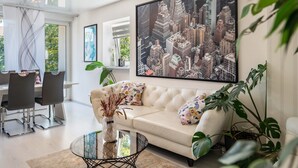 A modern living room with a white sofa, a glass coffee table, and a large cityscape painting on the wall.