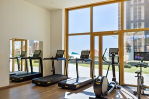 Ein Fitnessstudio mit Laufbändern, Crosstrainern und Fahrrädern, großen Fenstern und Blick auf Gebäude im Freien.