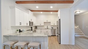A modern kitchen with white cabinetry, a marble island, wooden stools, and a staircase leading to another level. - Tabby Cottage: 3  BR, 3.5  BA House in Jekyll Island, Sleeps 6 (Jekyll Island)