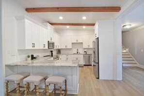 A modern kitchen with white cabinetry, a marble island, wooden stools, and a staircase leading to another level. - Tabby Cottage: 3  BR, 3.5  BA House in Jekyll Island, Sleeps 6 (Jekyll Island)