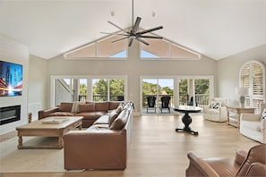 A spacious living room with a high ceiling fan, large windows, a leather sofa, a coffee table, and a TV. - Tabby Cottage: 3  BR, 3.5  BA House in Jekyll Island, Sleeps 6 (Jekyll Island)