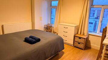 A bedroom with a bed, a chest of drawers, a small table, and a window with a view of buildings.