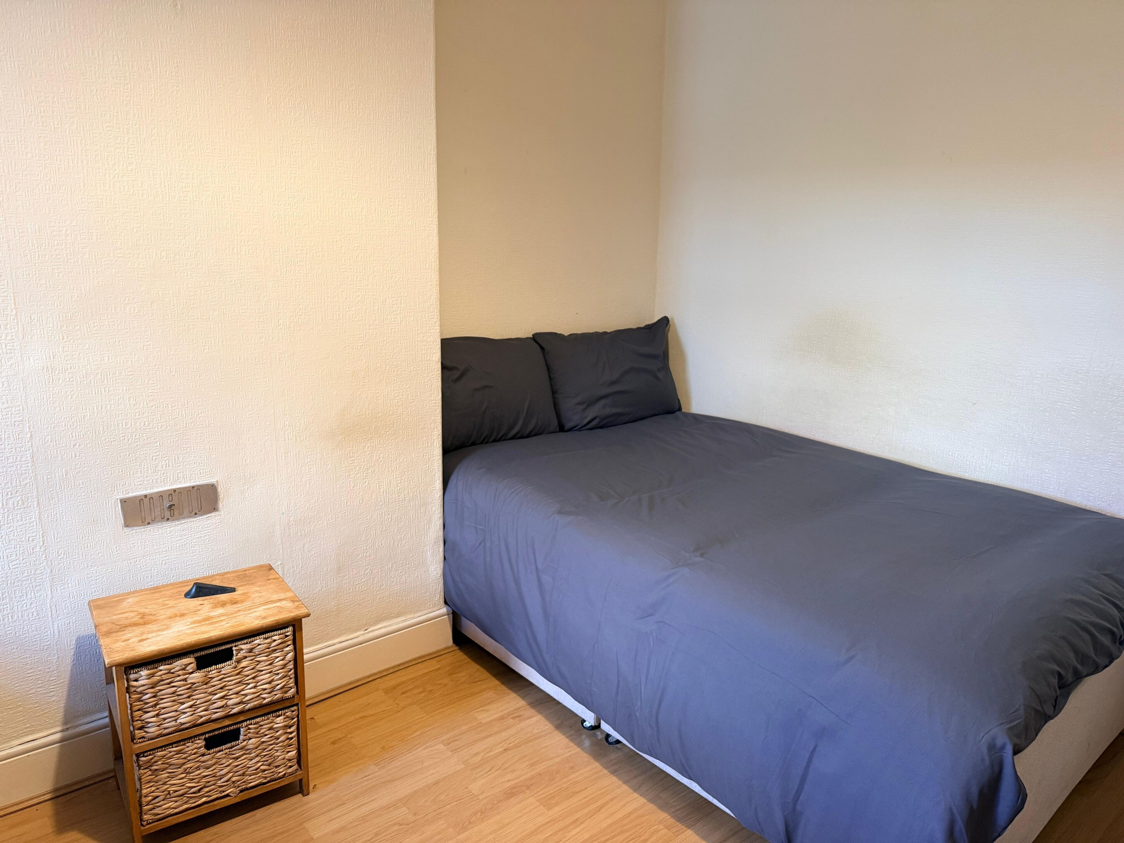 Ein Schlafzimmer mit einem Einzelbett, einem kleinen Nachttisch mit Körben und einer schlichten Wand.