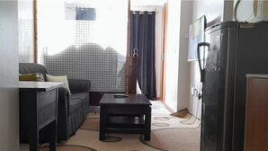 A compact living space with a sofa, a small table, a refrigerator, and a view through a window with a curtain. - 1-bed Apartment Sleeps 2 in Nairobi - Free Parking (Nairobi)