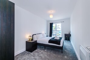 A modern bedroom with a bed, bedside table, lamp, and a view of the street through a window. - Modern and Stylish Flat - 1BDR (Liverpool)