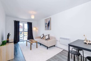 A modern living room with a sofa, a coffee table, a dining table with chairs, a large window with curtains, and a painting on the wall. - Stylish 1BDR Hospitals Nearby (Liverpool)