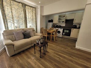 Ein Wohnzimmer mit einer Couch, einem Couchtisch und einem Essbereich mit Stühlen. Im Hintergrund sind eine Waschmaschine und Küchengeräte erkennbar.