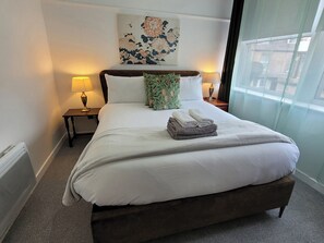 Ein ordentlich bezogenes Bett mit weißer Bettdecke, zwei Kissen und einem grünen Kissen mit Blumenmuster. Zwei Nachttischlampen, ein an der Wand hängendes Kunstwerk und ein Fenster mit Vorhängen sind ebenfalls zu sehen.